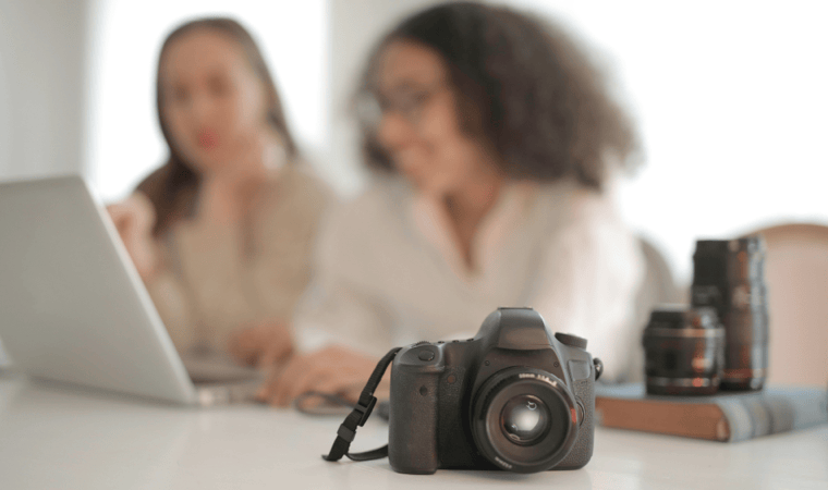 An SLR camera in the foreground with two women in front of a laptop blurred in the background