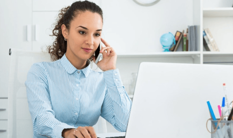 A professional young woman on her phone, sitting at a desk in front of a laptop