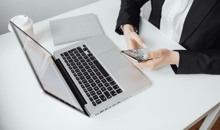 An overhead shot of a woman in a suit holding a phone, with a laptop, notebook, and coffee cup on a desk